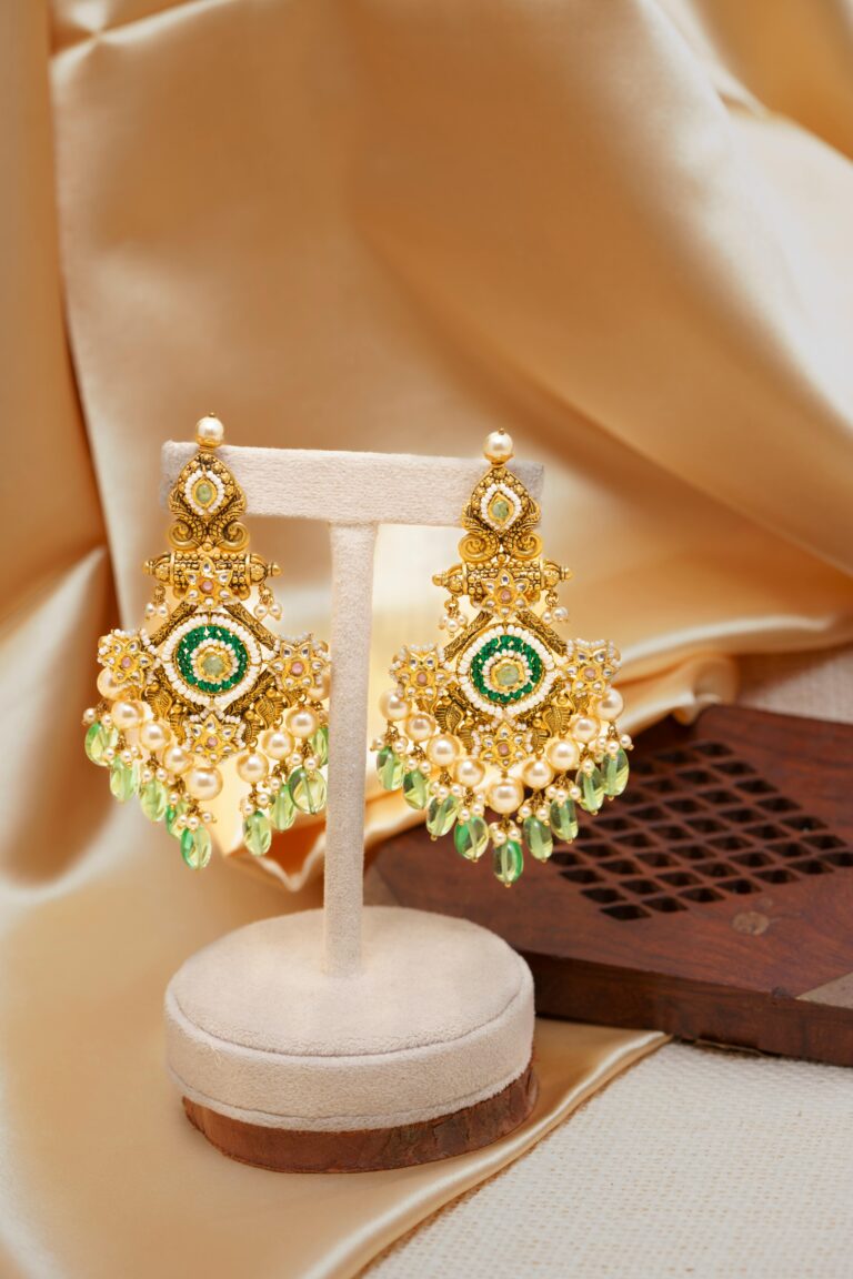 Home Close-up of luxurious gold chandelier earrings with green gems on a beige background.